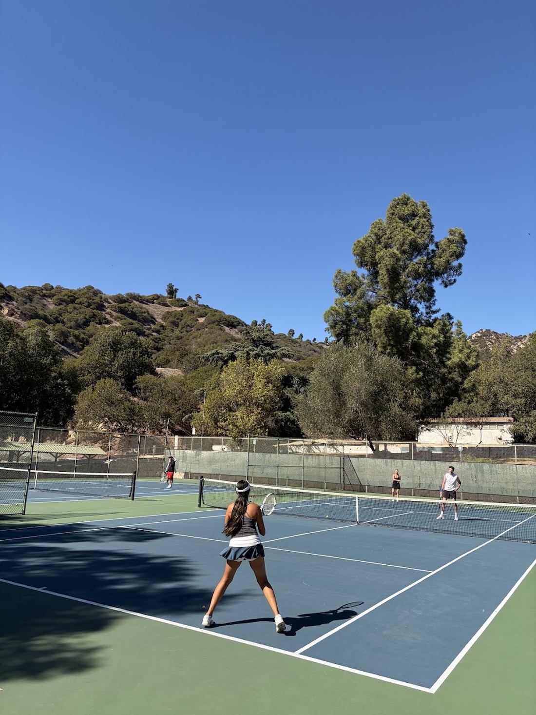 A player at the baseline waiting to return, courts busy behind her.